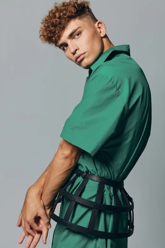 Young man with curly hair wearing green outfit and black harness posing against gray background in studio. Fashion model with serious expression and stylish clothing for editorial shoot.