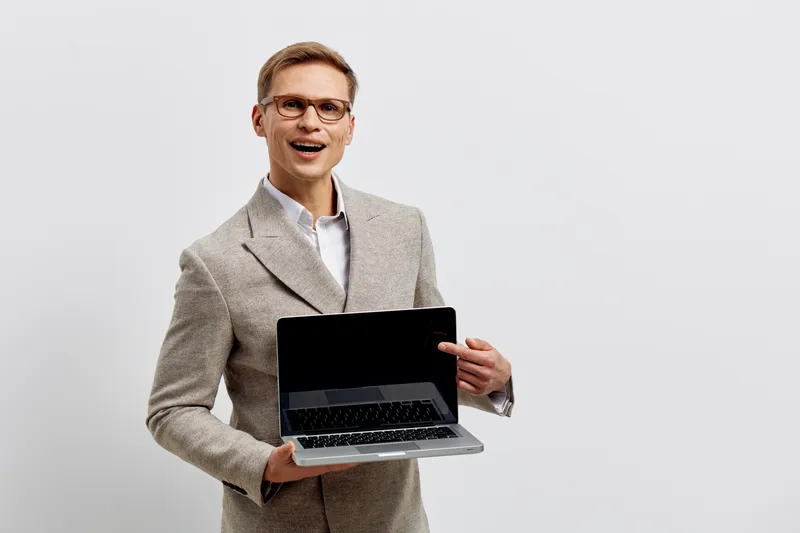 Smiling man with glasses wearing a gray blazer holding a laptop computer in front of a white background, professional business style, studio portrait, technology concept, modern office attire, happy e