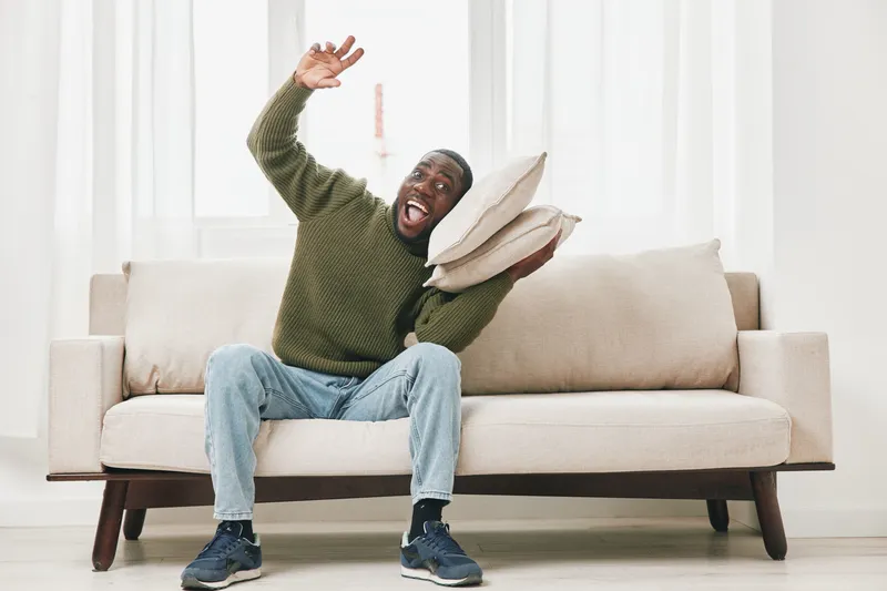 Joyful young man in olive green sweater sitting on a light couch, playfully holding pillows in a bright, airy room, capturing a moment of happiness and relaxation