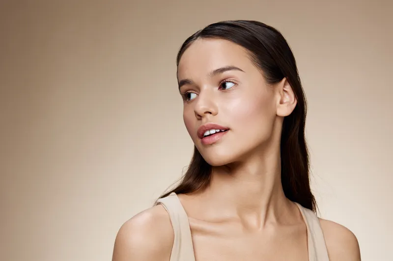 Young woman with long hair and natural makeup looking over her shoulder against a neutral beige background, portraying confidence and serenity in a beauty concept