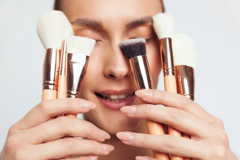 Close up of woman holding various makeup brushes near her face, soft focus on tools, natural skin tone, minimalistic background, beauty concept, cosmetic accessories.