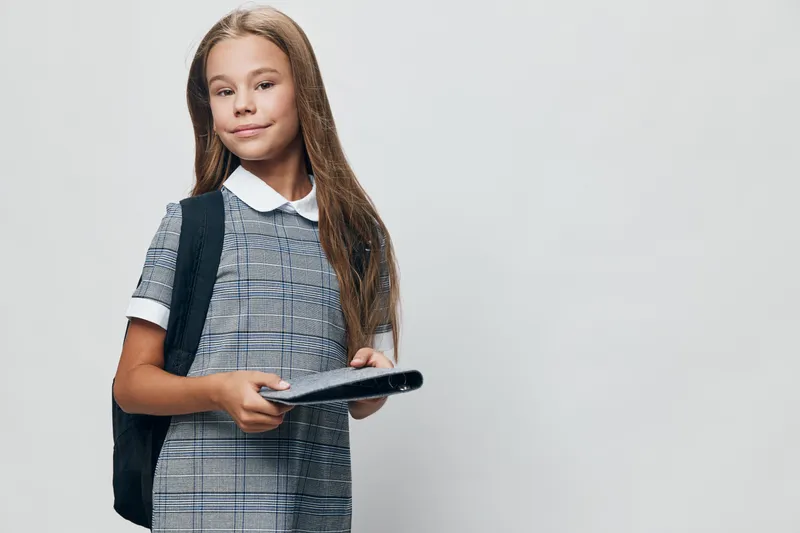 Schoolgirl with backpack holding notebook isolated on grey background standing and looking at camera with slight smile. Child education, student preparation and knowledge concept.