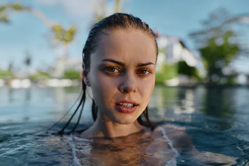 Cheerful woman in a sunlit pool, close up above water, wet hair and relaxed smile, shimmering reflections, outdoor leisure vibe, natural colors, serene and refreshing moment