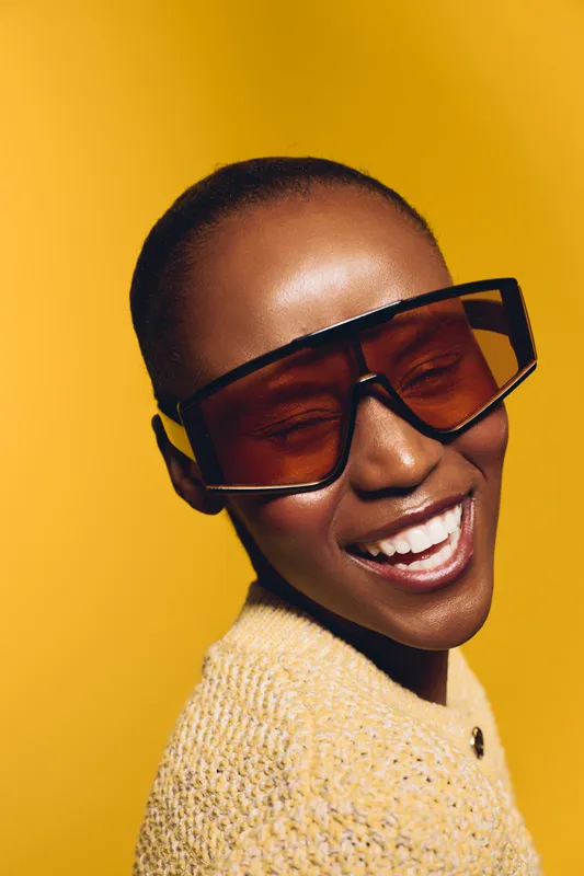 Smiling African woman with dark skin wearing oversized sunglasses and a textured yellow sweater against a bright yellow background. Concept of fashion, joy, and vibrant style.