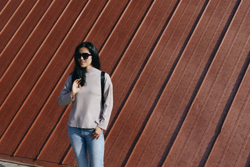 Woman wearing sunglasses with a backpack posing casually on an urban street, against a textured red wall, showcasing street fashion and outdoor style in daylight.