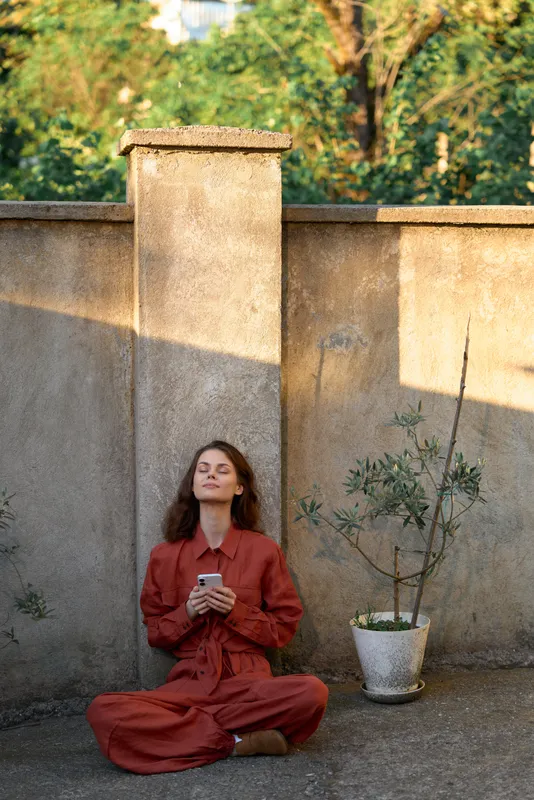Woman lifestyle outdoors, seated in a red dress, calm pose, eyes closed, holding a smartphone, warm sun, soft breeze, stone wall background, lush greenery nearby