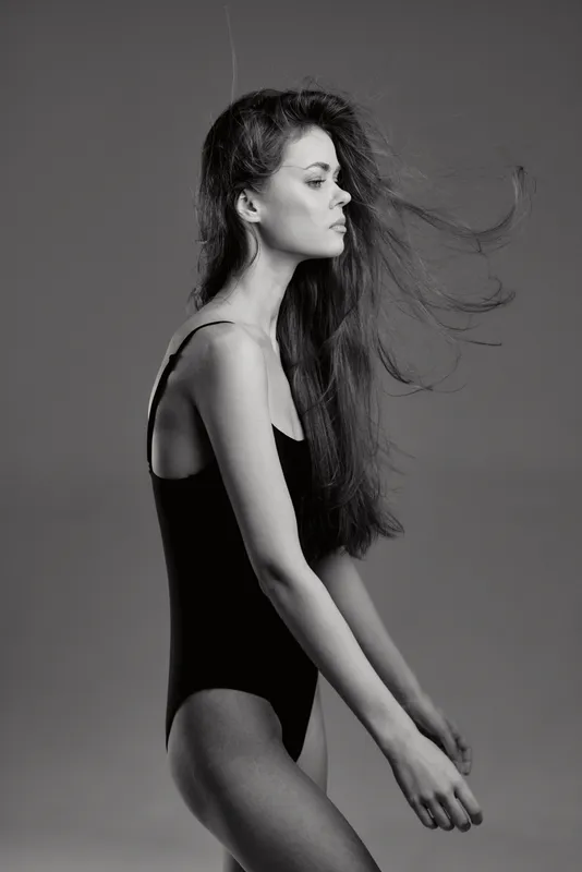 Black and white portrait of slender woman with flowing hair in minimalist outfit, showcasing elegance and grace in studio setting.