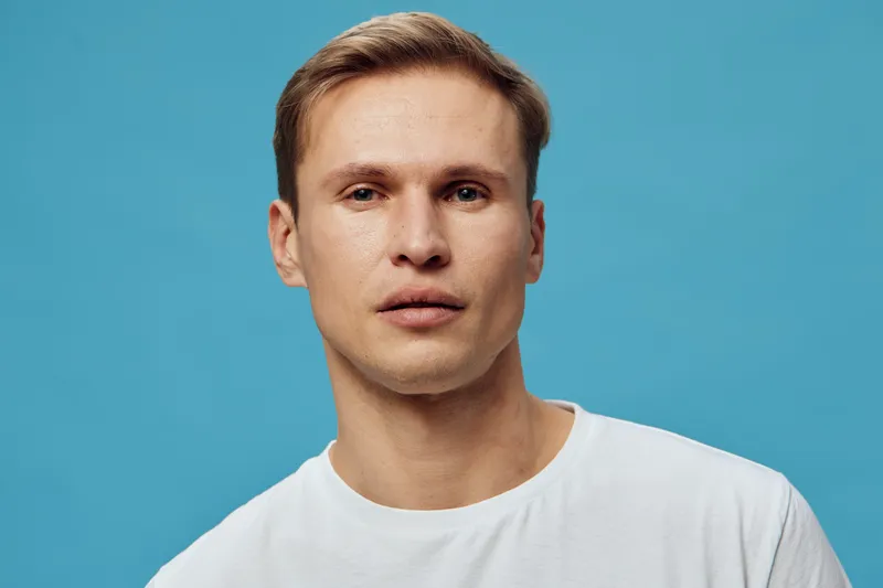 Close up serious young caucasian man wearing white t-shirt look camera isolated on light blue background mental health and gadgets concept portrait.