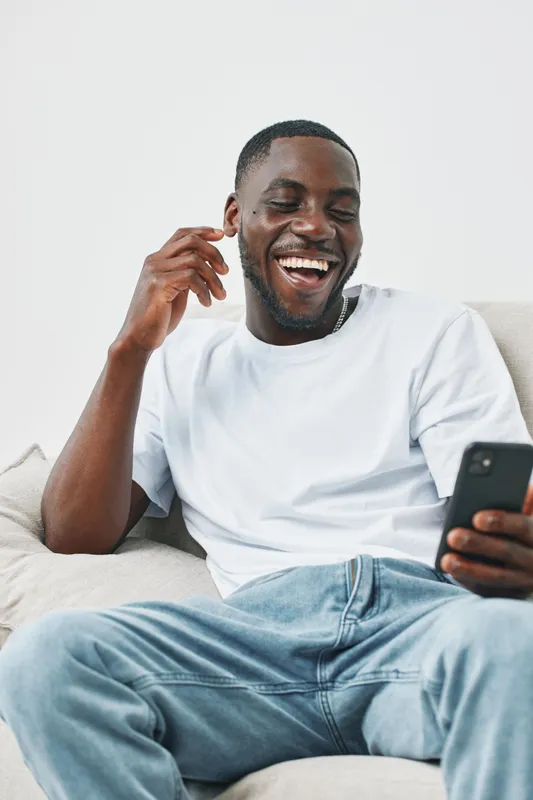 Happy young man laughing while using smartphone on a couch in casual outfit with bright background, conveying joy and relaxation