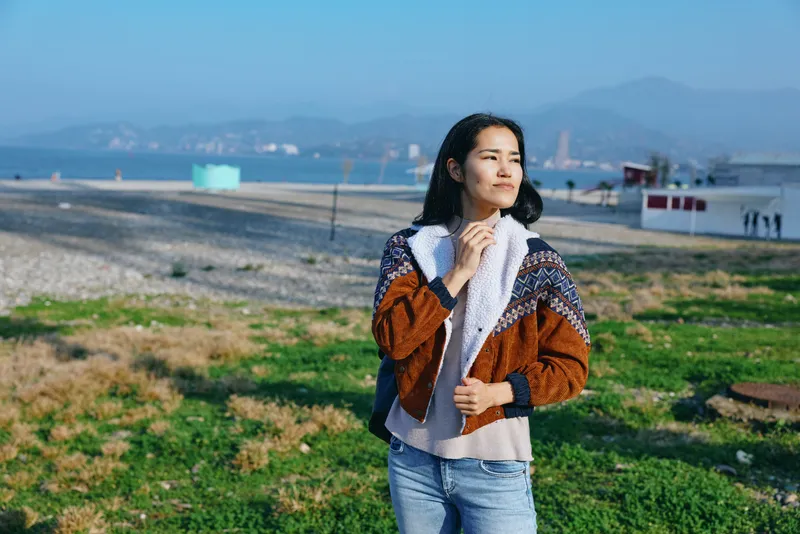 Outdoor scene with a woman in a patterned knit coat and scarf by the beach, enjoying a breezy day near calm sea and distant hills, embodying casual fashion and coastal mood