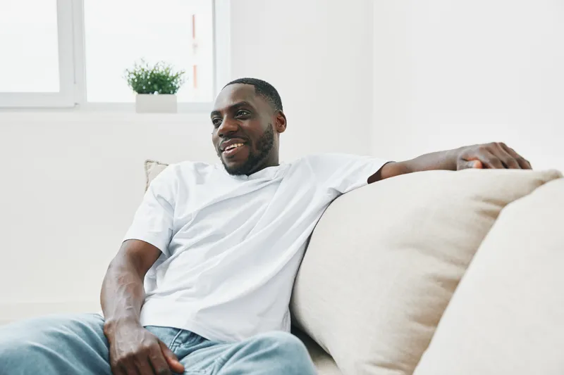 Happy young African American man relaxing on a sofa in a bright living room, wearing casual clothes and smiling, representing comfort and joy at home