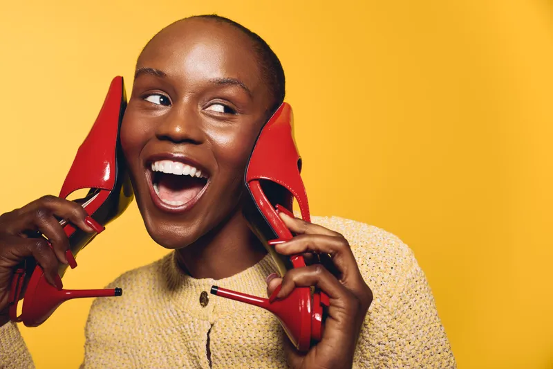Happy black woman with dark skin holding red high heel shoes near her ears smiling broadly on yellow background. Concept of fashion, joy, beauty, and playful mood in studio.