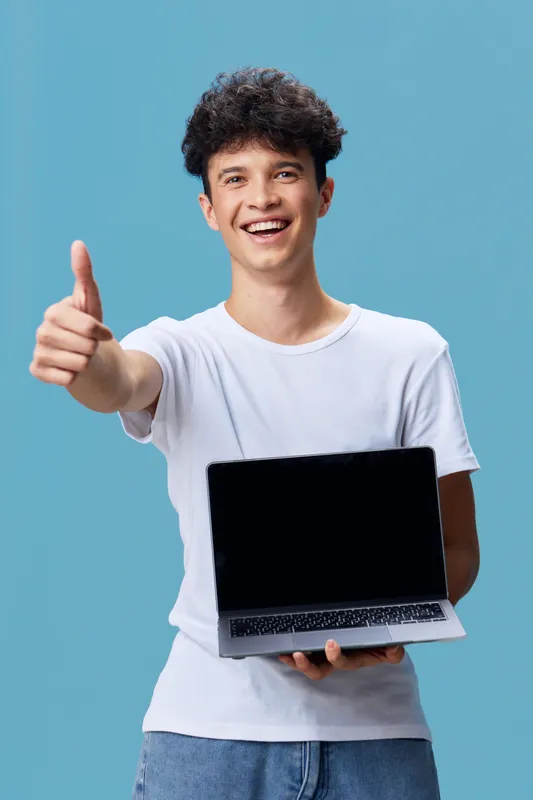 young man smiling showing laptop with blank screen and thumbs up gesture, wearing white t-shirt against blue background, positive expression and casual style