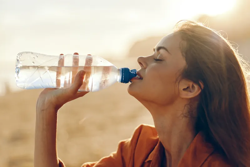 Woman drinking water from a bottle outdoors in warm sunlight, close up profile, soft blurred background, hydration wellness concept, summer thirst relief, active lifestyle peaceful mood