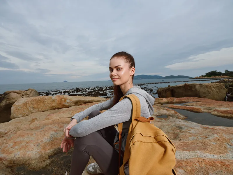 Young woman with a backpack sitting on rocks by the sea, wearing casual attire, smiling, with a cloudy sky background and calm ocean, perfect for lifestyle imagery