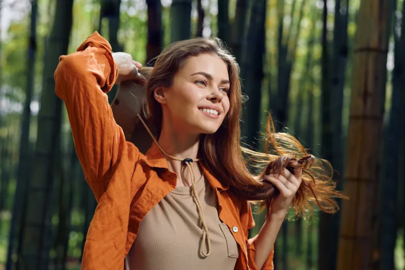 Woman portrait with smile in nature among bamboo outdoors, casual jacket and flowing hair, relaxed and cheerful in sunlit green forest for lifestyle and travel.