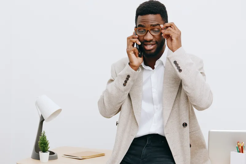 Smiling man in business casual outfit adjusting glasses and talking on mobile phone in modern office interior with white wall background. Happy, confident, professional male model representing busines