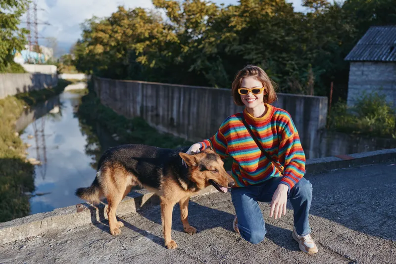 young woman kneeling by an urban canal with a german shepherd, wearing a colorful striped sweater and sunglasses, smiling in casual outfit during golden hour outdoors.