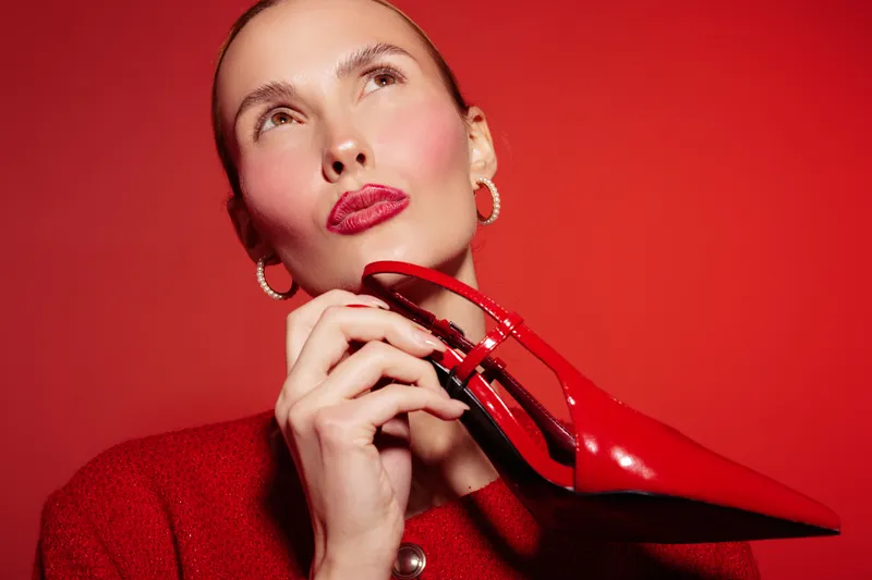 Stylish woman in red sweater holds shiny red high heel shoe near face on red background. Concept of fashion, elegance, beauty, and luxury accessories with vibrant color contrast.