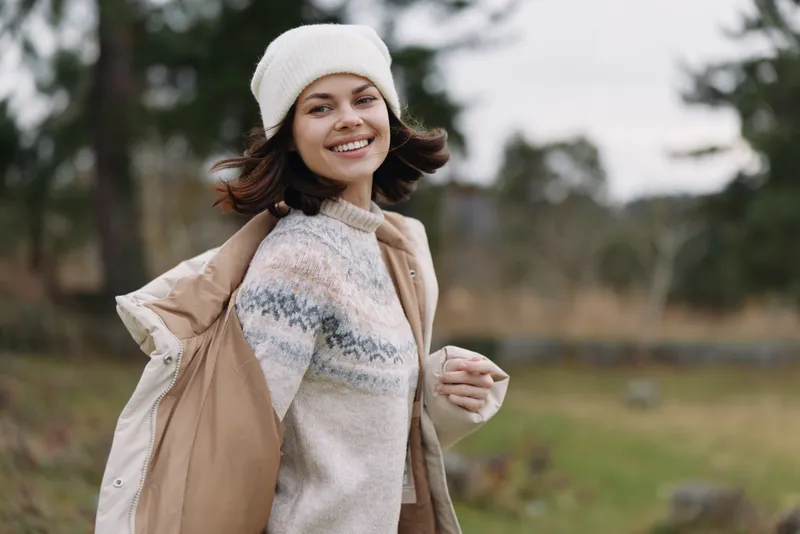 Woman in a cozy sweater and coat outdoors, smiling with a beanie, carrying a light jacket, autumn scenery and soft natural light frame a warm lifestyle portrait.