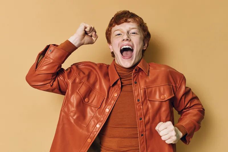 Excited young man with red hair wearing brown leather jacket and sweater cheering with raised fists on beige background. Expression of joy and celebration in casual outfit.