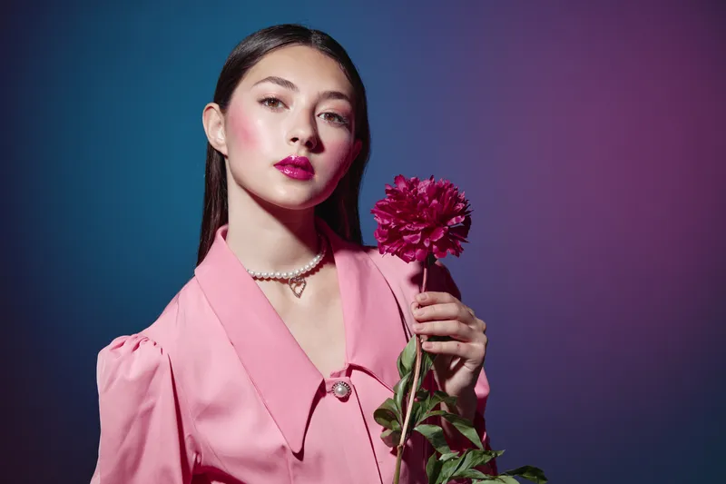 Young woman with pearl necklace wears pink outfit holding deep red carnation flower. Glossy magenta lips and natural makeup on blue purple gradient background. Beauty fashion concept.