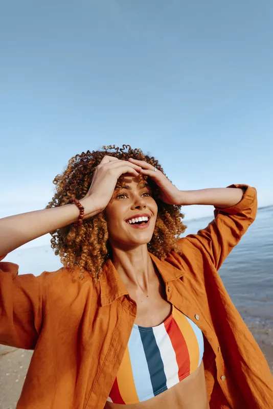 Young woman with curly hair smiles joyfully while standing on the beach wearing a colorful striped swimsuit and an orange shirt under a clear blue sky.
