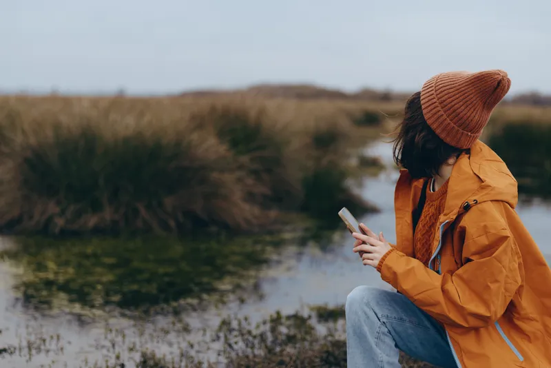 Woman lifestyle outdoors, sitting by river, wearing orange coat and knitted hat, reading on phone, autumn countryside nature scene, soft natural light, calm peaceful mood, blurred background