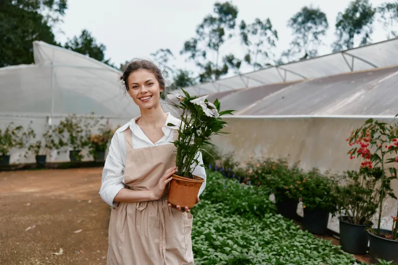 Young woman in apron holding potted plant in garden greenhouse, showcasing gardening passion and joy in a serene environment