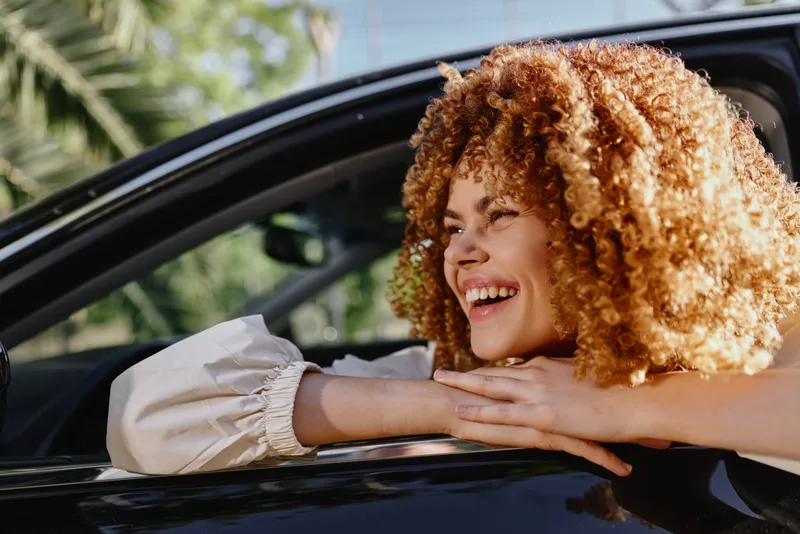 Smiling woman with curly hair leans out of car window, enjoying sunny day with tropical palm background. Bright outdoor scene exudes happiness and freedom.