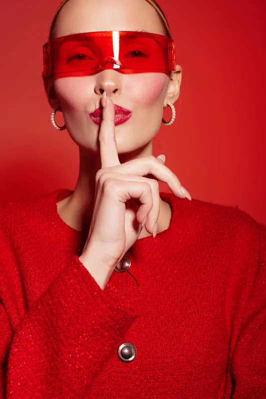 Stylish woman in red sweater and red sunglasses makes silence gesture with finger on lips. Bright red background and fashion accessories create bold modern look and secret concept.