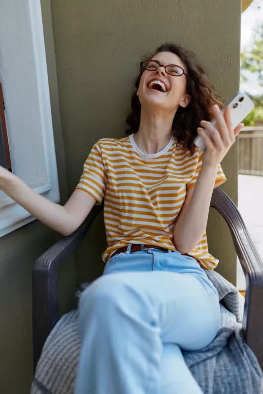 A woman with glasses laughs while using a smartphone on a sunny balcony, casual home setting. A cheerful moment of everyday lifestyle, relaxed and connected.