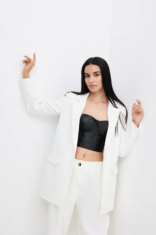Young woman in white suit jacket and trousers posing confidently against white corner wall, wearing black corset top and displaying modern fashion style.
