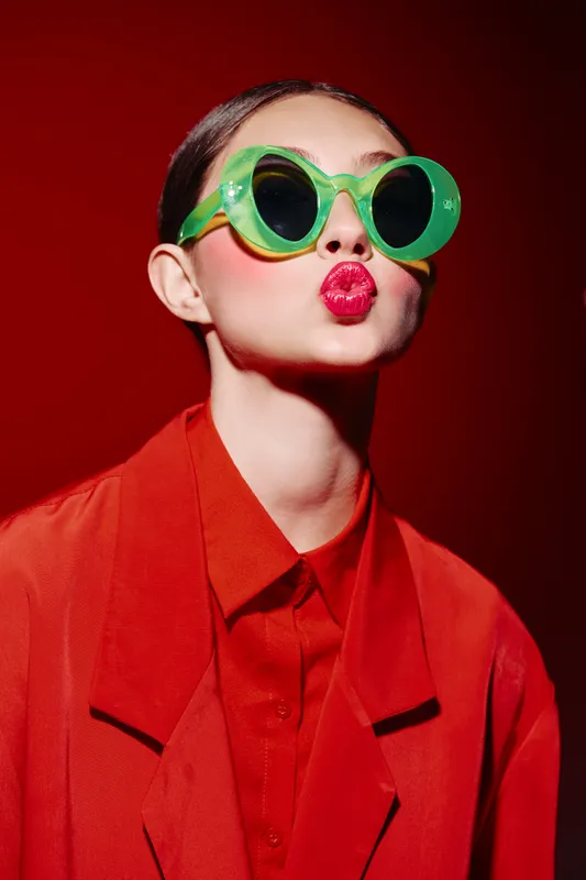 Young fashion woman with bright red lipstick wearing oversized green sunglasses and stylish red outfit on red background. Creative makeup, bold style, beauty concept.