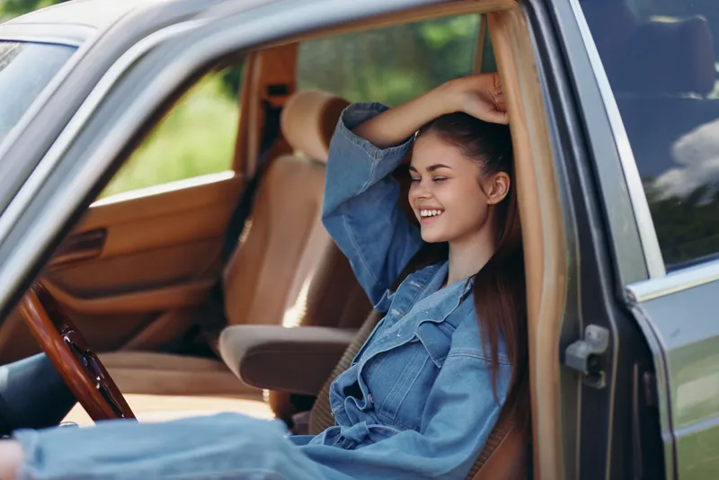 Happy young woman enjoying a summer day in a car, wearing denim attire and smiling, with a green natural background and warm sunlight.