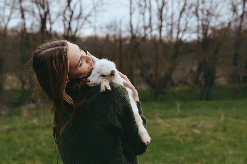 Warmhearted woman embraces a fluffy goat kid in an open meadow, showcasing rustic lifestyle and animal companionship. Natural daylight, soft colors, and gentle mood highlight wellbeing, bonding, and