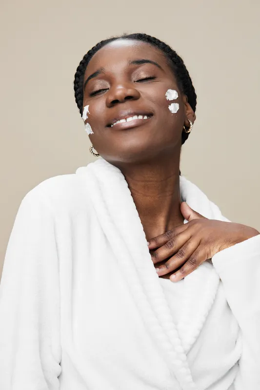 Young Black woman applying moisturizing cream on her face, radiating joy and confidence in a minimalist light beige background, promoting skincare and self care rituals