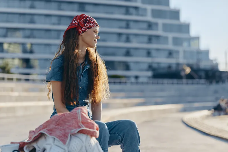 Lifestyle woman in modern boho-western denim street style sitting outdoors, wearing red bandana with natural sunlight and film color tones, casual urban fashion vibe.