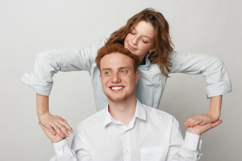 Happy young couple with red hair and curly hair smiling and holding hands in a playful pose on a light background. Concept of love, friendship, and positive emotions.