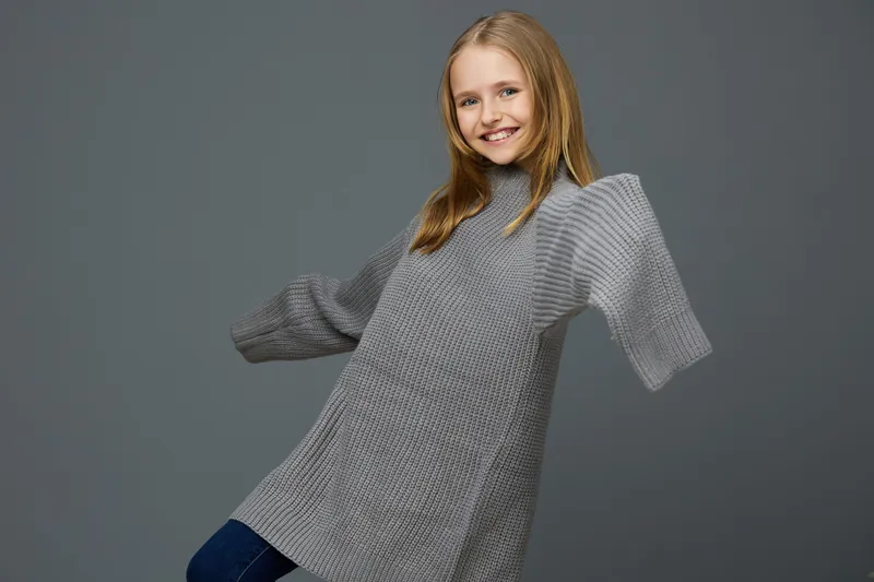 A cheerful young girl in a cozy gray sweater showcasing confident and playful emotions against a muted gray background, reflecting autumn fashion and winter warmth