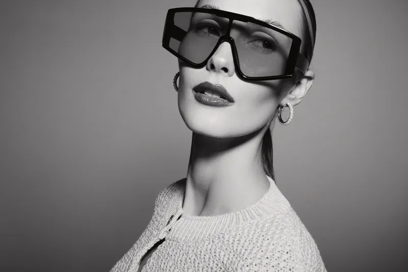 Stylish white woman with pale skin wearing large black oversized glasses and hoop earrings poses in studio with neutral background. Fashion concept.