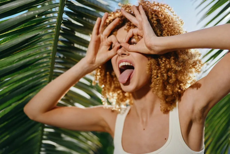 Playful woman with curly hair making funny glasses gesture by palm trees outdoors, expressing joy and summer vibes in bright natural light.