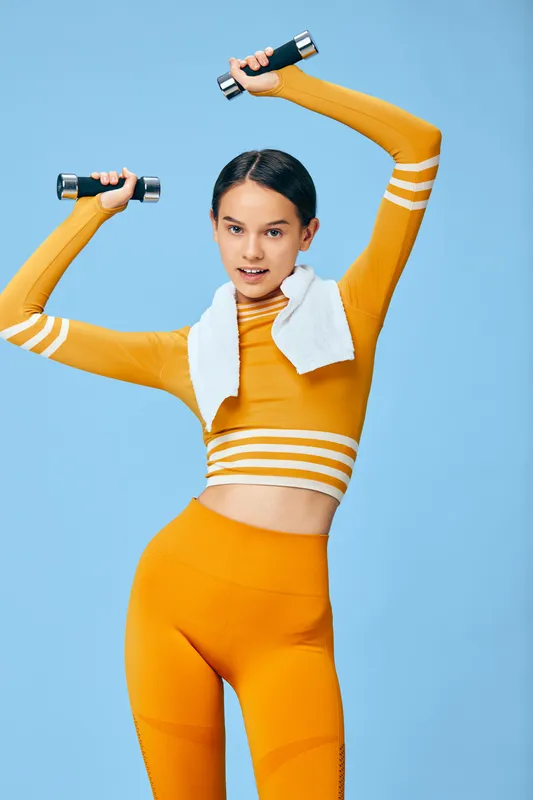 Fitness, sport, woman lifting dumbbells in bright orange athletic set with towel around neck, dynamic workout pose against blue backdrop, energetic training for strength and health.
