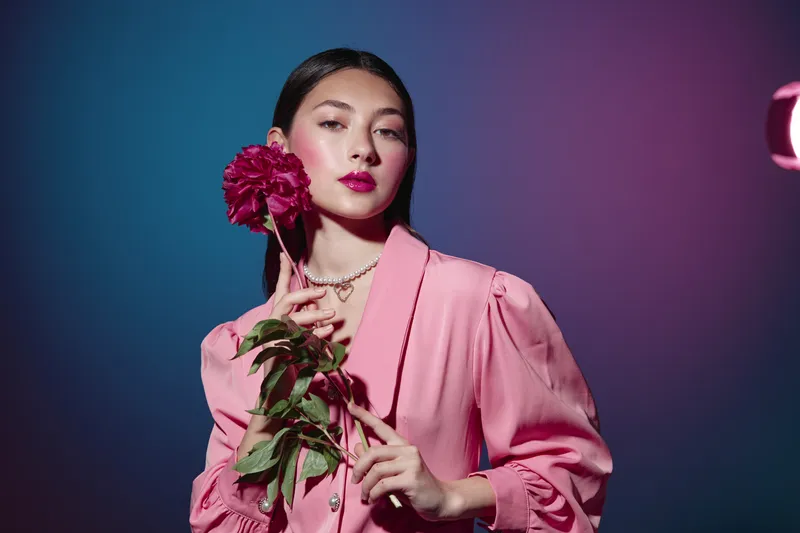 Young woman in a pink blazer holds dark red peony flower close to her face in a stylish beauty studio. Dramatic lighting highlights creative fashion and makeup concepts.