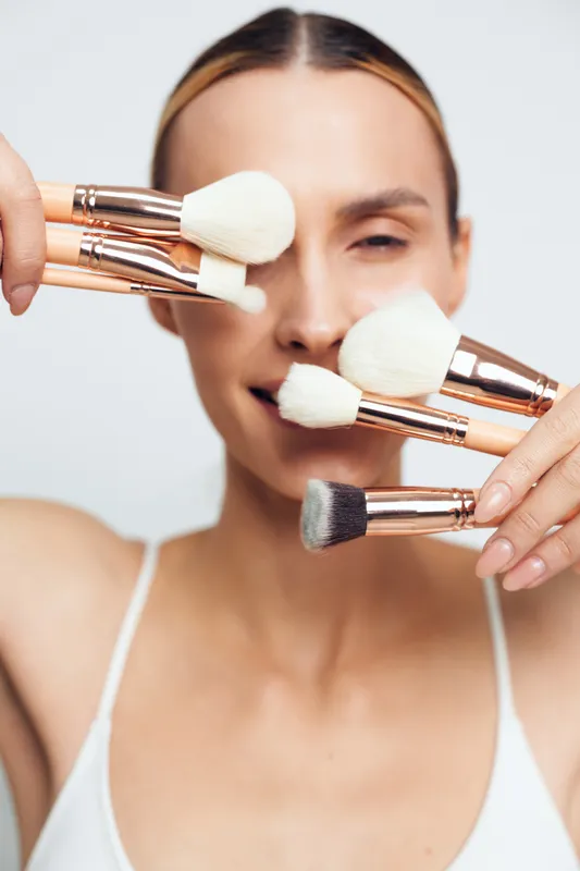 A smiling woman holds several makeup brushes with white bristles near her face against a plain background. The concept highlights beauty tools and skincare routine essentials.