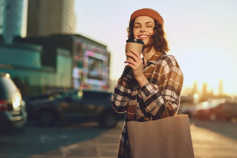 Woman with coffee and shopping bag smiles on an urban street at golden hour glow, candid lifestyle portrait showing authenticity, mindful living and emotional storytelling.