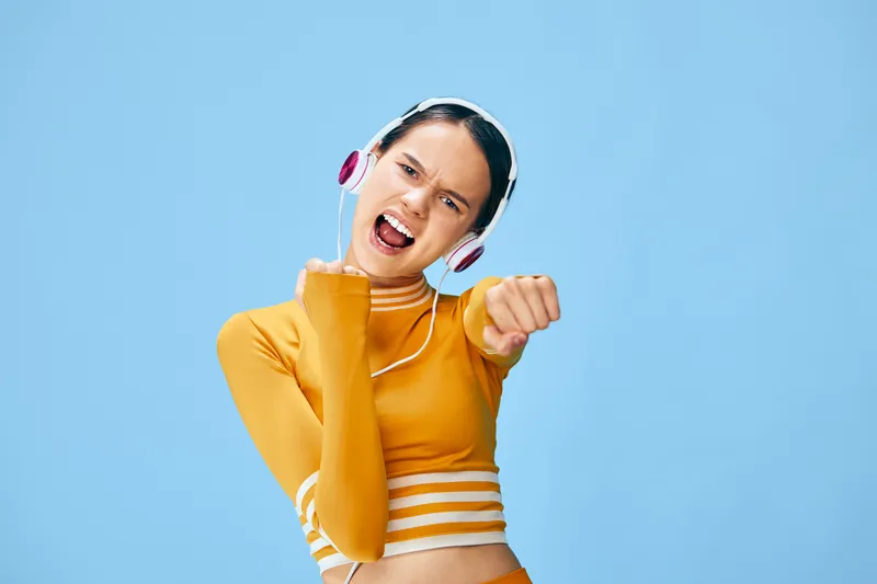 Woman in a sporty orange top with headphones, striking a dynamic pose against a vivid blue background, showcasing fitness energy and a stylish active lifestyle.