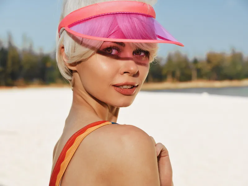 Portrait of a stylish woman wearing a pink visor on a sunlit beach, highlighting smooth skin, confident gaze, and a breezy summer mood under clear skies.