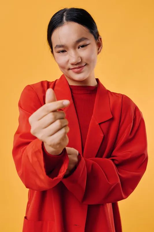 Young woman in red blazer making finger heart gesture with smile standing against orange background. Asian female expressing love and positivity with casual confident posture.