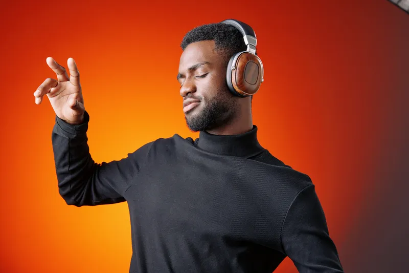 Young Black man enjoying music with headphones, expressing joy in a vibrant orange background, wearing a stylish black turtleneck, perfect for modern lifestyle concepts